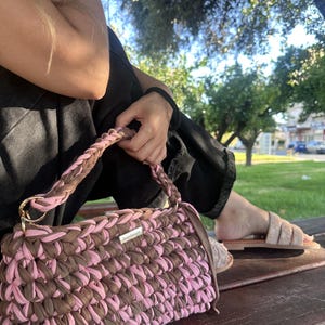 Puede incluir: Un bolso de mano rectangular de ganchillo con un patrón tejido de tiras de tela alternadas en marrón y rosa claro. El bolso incluye un asa trenzada a juego, anillas de metal dorado y una pequeña etiqueta rectangular con la palabra handmade.