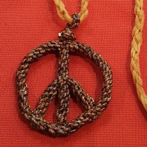 Puede incluir: Un colgante con el símbolo de la paz tejido en marrón sobre un cordón beige. El colgante está hecho de material tejido apretado, y el cordón está anudado en la parte superior. El fondo es de color rojo liso.