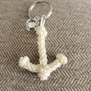 May include: Cream-colored, nautical-themed keychain featuring a woven anchor design. The anchor is attached to a silver keyring with a small crab charm. The keychain is made of a soft, rope-like material.