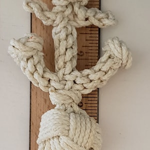 Puede incluir: Ancla de temática náutica hecha de cuerda blanquecina, de aproximadamente 13 cm de altura. El ancla está intrincadamente anudada, con una base redondeada y un diseño de barra transversal. Está colocada junto a una regla de madera.