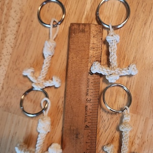 May include: Three handmade anchor keychains made of off-white rope, each attached to a silver keyring. A wooden ruler is placed between the keychains. The keychains are nautical themed.