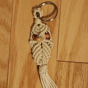 Puede incluir: Llavero de macramé color crema con un diseño decorativo, con cuentas de madera y una borla. El llavero está unido a un anillo de metal plateado, ideal para llaves o dijes de bolso.