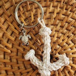 May include: A silver-colored keychain with a small turtle charm and a white rope anchor design. The keychain is attached to a large silver ring. The background is a woven, light brown surface.