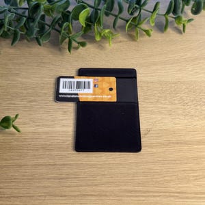 May include: A rectangular black card holder with a textured surface. A small orange and white tag is inserted into the top, featuring a barcode, a circular hole, and the printed text www.hamiltonbindingcentres.co.uk.