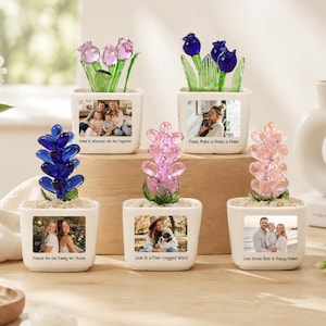 May include: Five white square ceramic planters featuring custom photo prints and text, each containing a glass flower sculpture. The sculptures include blue and pink heart-shaped glass petals and green glass stems set in white pebbles.
