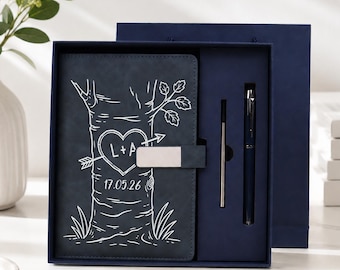 Può includere: Un set di taccuini blu scuro con un'illustrazione a linee bianche di un tronco d'albero con un cuore, una freccia, le lettere L+A e la data 17.05.26. Il set include una penna blu abbinata e una ricarica all'interno di una scatola regalo.