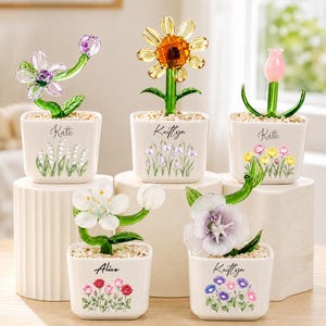 May include: Five square white ceramic planters containing glass flowers. Each planter features a name, including Kate, Kaitlyn, and Alice, and floral illustrations. The glass flowers include a purple blossom, a yellow sunflower, a pink tulip, a white flower, and a large purple bloom, all set in small pebbles.