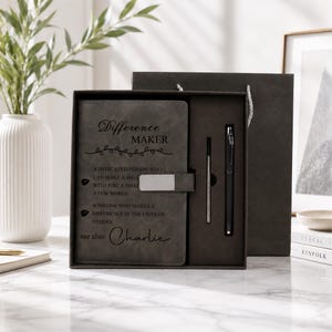 Pu&ograve; includere: Un set regalo composto da un taccuino grigio scuro e una penna in una scatola coordinata. La copertina del taccuino riporta il testo Difference MAKER, una definizione del termine e il nome Charlie. Una chiusura in metallo argentato fissa il taccuino e include una penna nera.