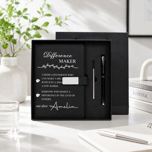 Pu&ograve; includere: Un set regalo composto da un taccuino nero e una penna in una scatola coordinata. La copertina del taccuino riporta il testo "Difference Maker" seguito da una definizione e dal nome "Amelia" in corsivo bianco. Una chiusura in metallo argentato fissa la copertina.