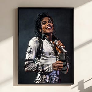 May include: A framed portrait of a person with dark curly hair, wearing a metallic silver long-sleeved shirt with black straps and buckles, holding a silver microphone. The person is smiling, set against a dark background within a black frame.