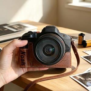 May include: A black digital camera featuring a brown textured leather case with the name Noah embossed on the side. The camera includes a matching brown leather neck strap, a circular lens, and a black body with visible buttons and dials on top.