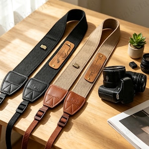 May include: Two camera straps, one black and one beige, featuring a herringbone weave pattern. Each strap includes a brown leather patch with a camera icon and text, a small metal camera charm, and leather end tabs with adjustable nylon webbing.