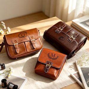 May include: Three brown leather-style satchel bags with brass-toned buckles. One bag features a camera icon and the text "Luna's photography," another has a camera icon, and the third displays a laurel wreath with the letter "M" and "Mia."