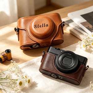 May include: A tan leather camera case with the name Stella embossed on the front lens cover, positioned next to a black camera housed in a dark brown leather case, a roll of 35mm film, and small white flowers on a light-colored surface.