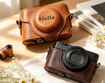 May include: A tan leather camera case with the name Stella embossed on the front lens cover, positioned next to a black camera housed in a dark brown leather case, a roll of 35mm film, and small white flowers on a light-colored surface.
