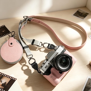 May include: A silver and black mirrorless camera encased in a pink leather-like protective cover with the name Violet printed on the front. It features a multi-textured camera strap in pink and grey with metal hardware and a small round pink pouch.