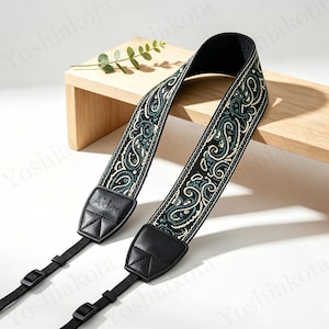 May include: A camera strap featuring a black fabric band with a teal and cream paisley pattern. The strap includes black leather end tabs with the name Aiden embossed on one side, black nylon webbing, and adjustable plastic buckles for length control.