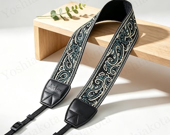 May include: A camera strap featuring a black fabric band with a teal and cream paisley pattern. The strap includes black leather end tabs with the name Aiden embossed on one side, black nylon webbing, and adjustable plastic buckles for length control.
