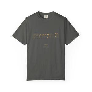 May include: A charcoal gray short-sleeve t-shirt featuring a gold-colored musical staff with notes and the text "UPBEAT" printed in gold. The shirt has a crew neckline and a standard fit, with a small brand label visible at the inner collar.