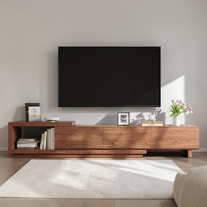 May include: A long, low-profile wooden media console with a natural wood grain finish, featuring an open shelf on the left side containing books and a decorative bowl, and three closed storage compartments. A flat-screen television is mounted above.