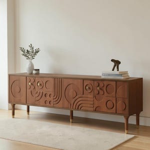 May include: A long, rectangular wooden sideboard featuring a medium-brown finish and intricate geometric relief patterns on its cabinet doors. The piece is supported by four tapered legs with gold-toned metal caps and includes small, circular gold-toned knobs.