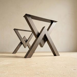 Puede incluir: Un par de patas de mesa de metal gris oscuro con un diseño geométrico y soportes en ángulo que forman una V central. Las patas tienen una placa de montaje rectangular plana en la parte superior con orificios pretaladrados para fijación.