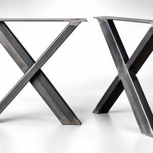 Può includere: Una coppia di gambe per tavolo a forma di X realizzate in metallo grezzo grigio scuro. Ogni gamba presenta una piastra di montaggio rettangolare piatta sulla parte superiore con fori preforati, collegata a due travi inclinate a forma di X.