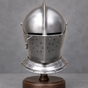 May include: A polished steel sallet helmet featuring a central ridge, a horizontal eye slit, and a visor with multiple circular ventilation holes. The helmet is secured to a circular dark brown wooden base with a series of small metal rivets.