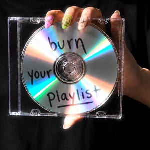 Könnte beinhalten: Eine transparente CD-Hülle aus Kunststoff mit einer reflektierenden silbernen Compact Disc. Auf der Oberfläche der Disc befindet sich der handgeschriebene schwarze Text "burn your playlist", der einen regenbogenfarbenen Lichtbrechungseffekt auf der metallischen Oberfläche erzeugt.