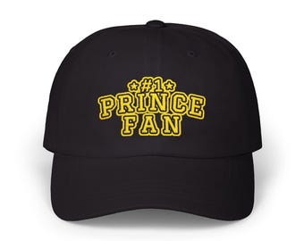 May include: A black baseball cap featuring yellow embroidered text that reads #1 PRINCE FAN, with a star on each side of the #1. The cap has a structured crown, a curved brim, and a button on the top center of the fabric dome.