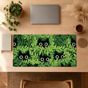 Op de afbeelding: Een rechthoekige bureaumat met een herhalend patroon van zwarte kattengezichten met grote witte cirkelvormige ogen die achter dicht groen gebladerte en bladeren vandaan kijken. Het ontwerp bedekt het gehele oppervlak van de mat in levendig groen.