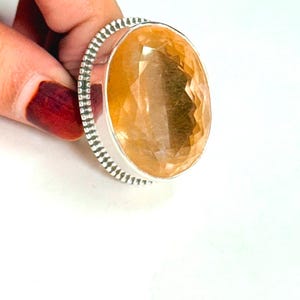 May include: A silver ring featuring a large, oval-cut, translucent yellow gemstone. The setting has a decorative, textured border with a series of small, vertical indentations surrounding the central stone, creating a distinct metallic frame.