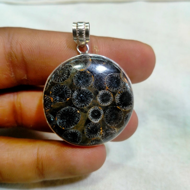 May include: A silver pendant featuring a round, black and white fossilized coral stone with a detailed pattern. The pendant is on a silver chain.