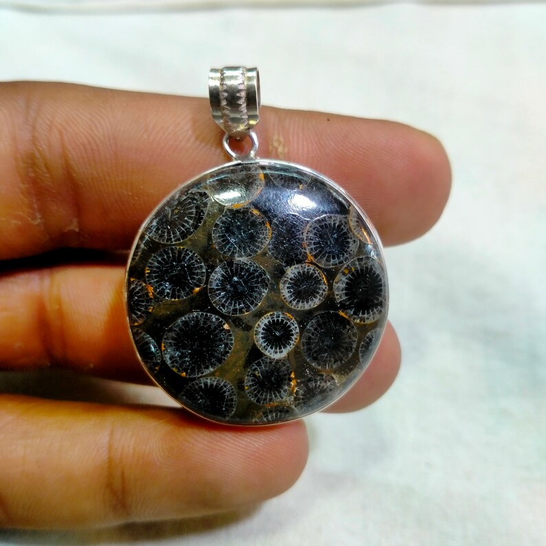 May include: A silver pendant featuring a round, black and white fossilized stone with a circular pattern. The stone is set in a silver frame and has a silver bail.