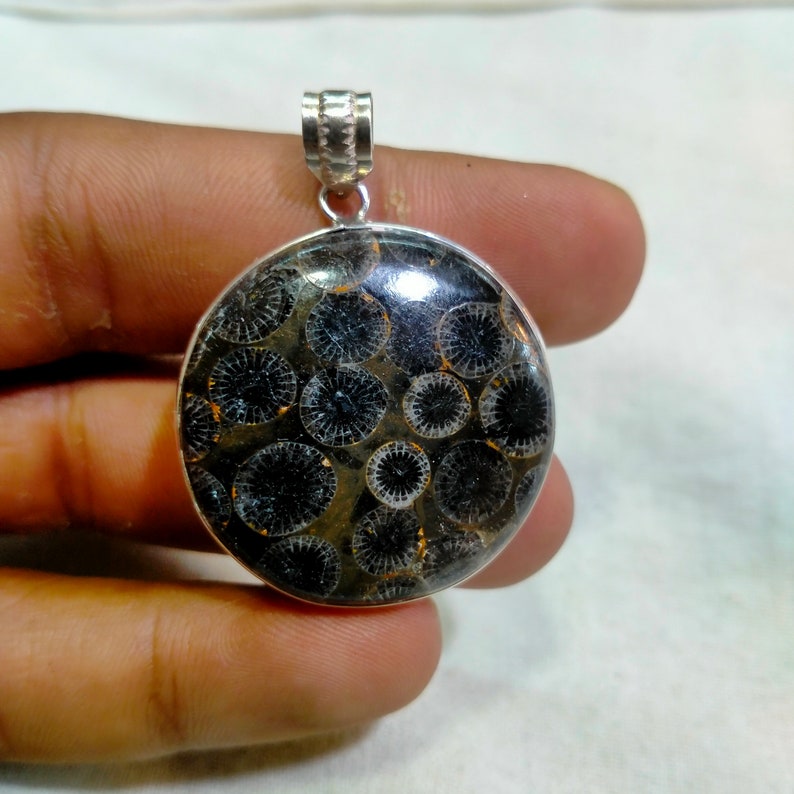 May include: A silver pendant featuring a round, black and white fossilized coral stone. The stone has a circular pattern of small, white circles within larger, black circles.