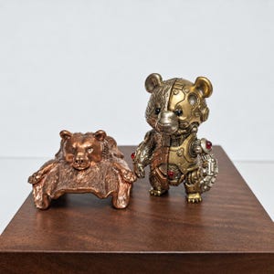 May include: A copper-coloured bear figurine in a prone position next to a standing mechanical bear figurine. The mechanical bear features a metallic gold and bronze finish with visible gear-like details, circular joints, and small red circular accents.