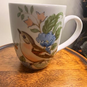May include: A white ceramic mug featuring a painted illustration of a small brown and tan bird perched among green leaves, pink bell-shaped flowers, and blue blossoms. The mug has a curved handle and a smooth, glossy finish.
