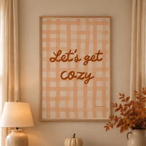 Peut inclure: Une affiche murale rectangulaire dans un cadre en bois clair présentant un motif à carreaux pêche et blanc avec le texte marron Let's get cozy centré sur l'impression. L'œuvre est exposée sur un mur au-dessus d'une console en bois.