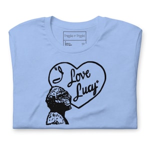 May include: Light blue t-shirt featuring a black and white illustration of a gorilla and a heart-shaped design with the words "I Love Lucy". The shirt has a crew neck and a tag that says "Praggles & Friggles".