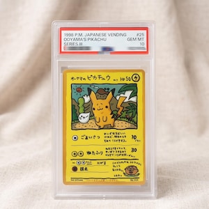 Puede incluir: Una carta coleccionable Ooyama's Pikachu de la serie japonesa Vending Series III de 1998, en un estuche de plástico transparente. La carta amarilla muestra una ilustración estilo dibujo a mano, texto en japonés, 50 HP y al artista Ooyama.