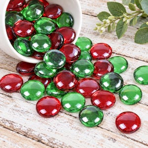 May include: A collection of smooth, round, translucent glass gems in vibrant red and green colors. Many of the polished glass stones are spilling out from a white ceramic bowl, while others are scattered across the surface.