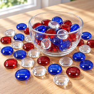 May include: A clear glass bowl filled with a collection of smooth, round, flat glass gems in red, blue, and clear colors. Additional matching glass gems are scattered around the base of the bowl, displaying a mix of translucent and solid tones.