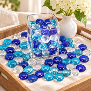 May include: A clear glass rectangular vase filled with smooth, round glass pebbles in shades of deep blue, light turquoise, and transparent clear. Additional matching glass pebbles are scattered across the surface around the base of the vase.
