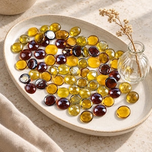 May include: A collection of round, flat glass gems in shades of amber, gold, and dark brown scattered across an oval, speckled ceramic tray. A small, clear, ribbed glass vase containing a single dried floral stem sits on the tray.