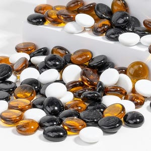 May include: A collection of smooth, rounded glass stones in a mix of solid white, translucent amber, and opaque black colors. The stones are scattered in a pile and arranged on tiered white surfaces, featuring a polished, reflective finish.