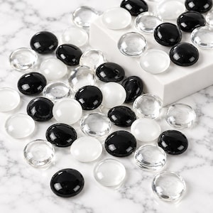 May include: A collection of smooth, round, flat-bottomed glass stones in solid black, opaque white, and transparent clear finishes. These decorative pebbles are scattered across a surface, with some resting on a white rectangular block.