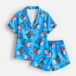 May include: A bright blue pyjama set featuring a short-sleeved button-down shirt and matching shorts. The fabric is covered in a repeating pattern of Thomas the Tank Engine, with white piping trim along the collar, front placket, and sleeve cuffs.