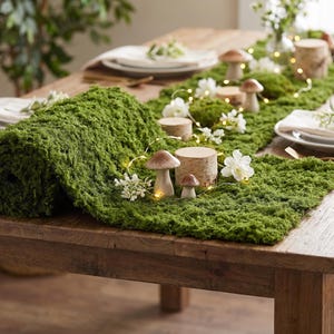 May include: A green moss table runner featuring a rolled end, decorated with small ceramic mushrooms, cylindrical wooden blocks, white artificial flowers, and a thin string of warm white fairy lights spread across the textured surface.