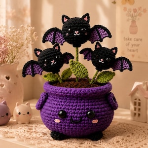 Pode incluir: Um vaso de crochê roxo em forma de caldeirão com um rosto sorridente, contendo três criaturas pretas de crochê semelhantes a morcegos em hastes verdes com folhas. O caldeirão tem pés e alças pretas e asas com detalhes em roxo.