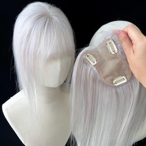 以下が含まれることがあります： ストレートの毛束と前髪があるプラチナホワイトのヘアトッパー。マネキンの頭部に装着されています。ヘアピースの裏側はベージュのベースになっており、頭皮にしっかりと固定するための3つの金属製クリップが付いています。